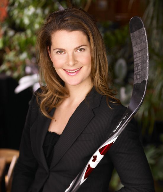 Jennifer Botterill. Headshot of Jennifer Botterill wearing a black jacket over a black top. She is smiling and holding a hockey stick. She is posing outside and the background blurry.
