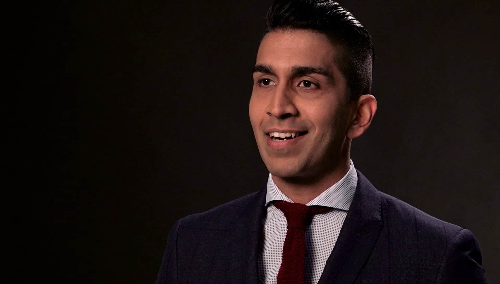 Nabil Karim. Headshot of Nabil Karim smiling, wearing a black suit and tie, posing in front a black background.