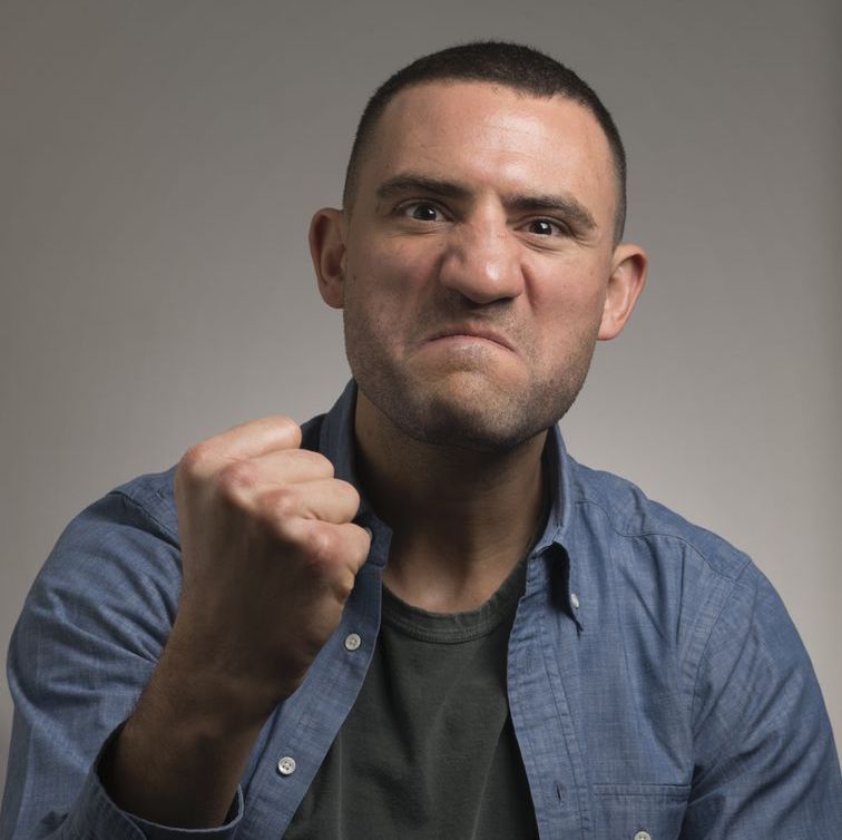 Paul Bissonnette. Headshot of Paul Bissonnette wearing a blue collared shirt over a black T-shirt. He is posing in front of a grey gradient background and holding up his fist.