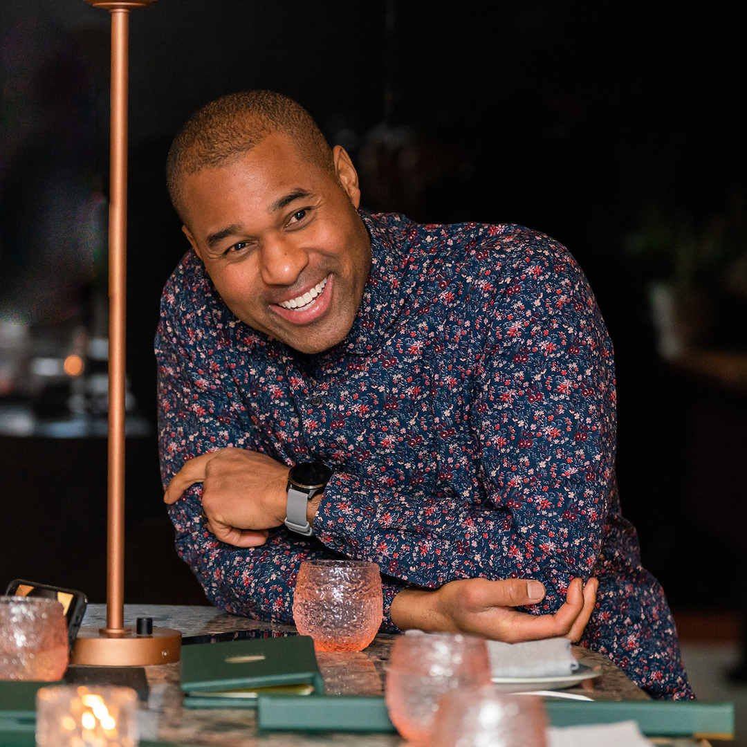 Cabral "Cabbie" Richards wearing a floral patterned shirt. He is smiling and leaning on his elbows on a table.