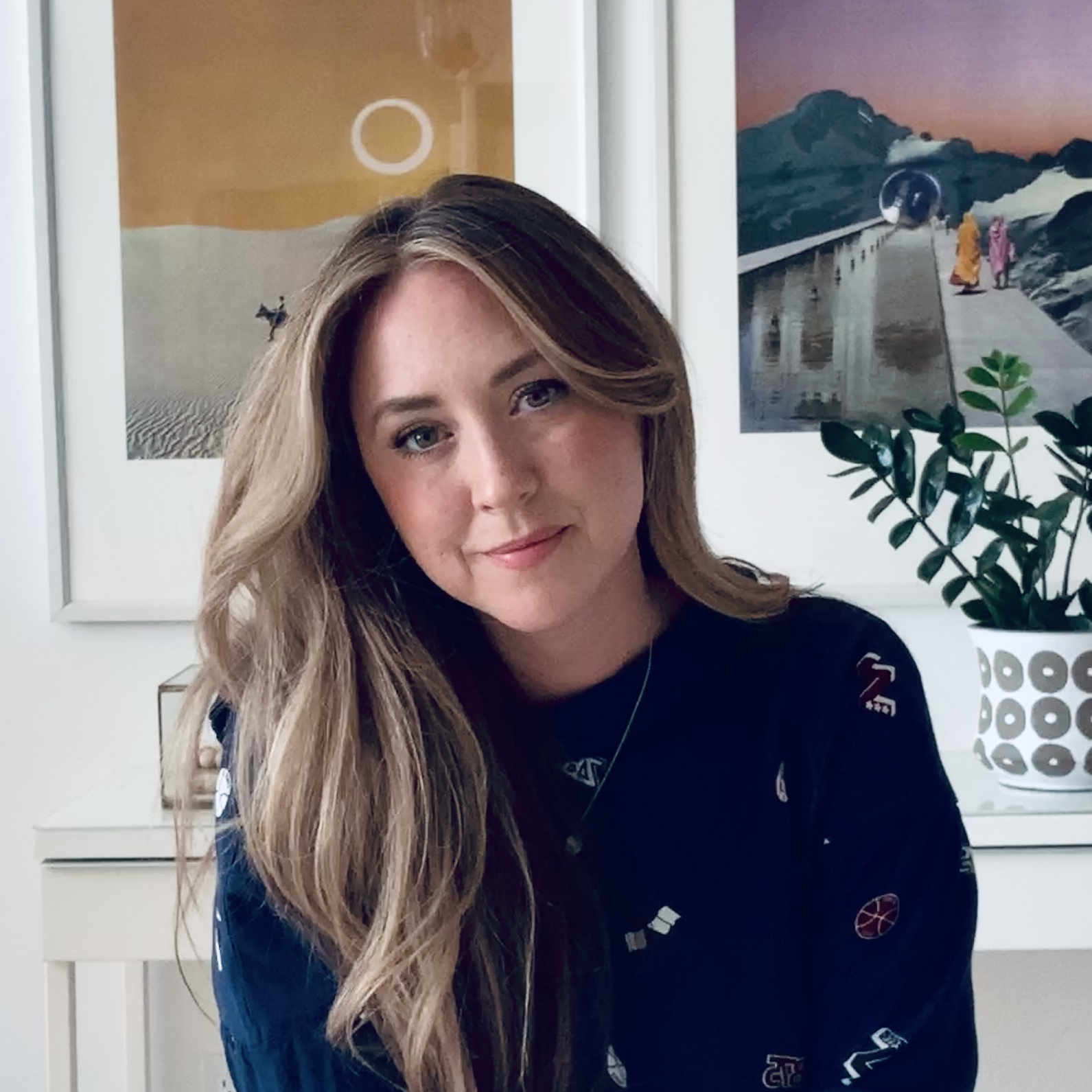 Chantel Chapman. Headshot or Chantel Chapman wearing a blue sweater. She is leaning on a white table in front of two framed art pieces.