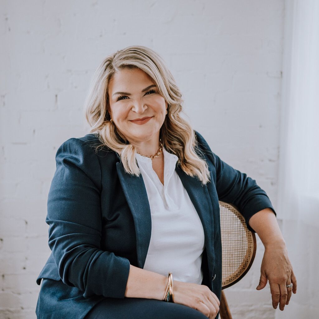 headshot of Canadian keynote speaker, Dr. Jody Carrington, sitting on a chair, wearing a white collared shirt and a navy blazer on top.