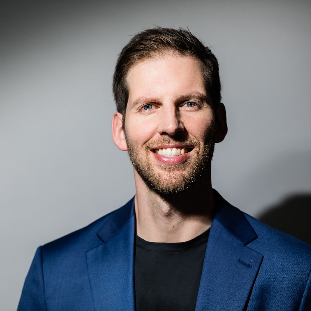 Jeremiah Brown. Headshot of Jeremiah Brown wearing a blue suit jacket over a black t-shirt. He is smiling and posing in front of a grey background.