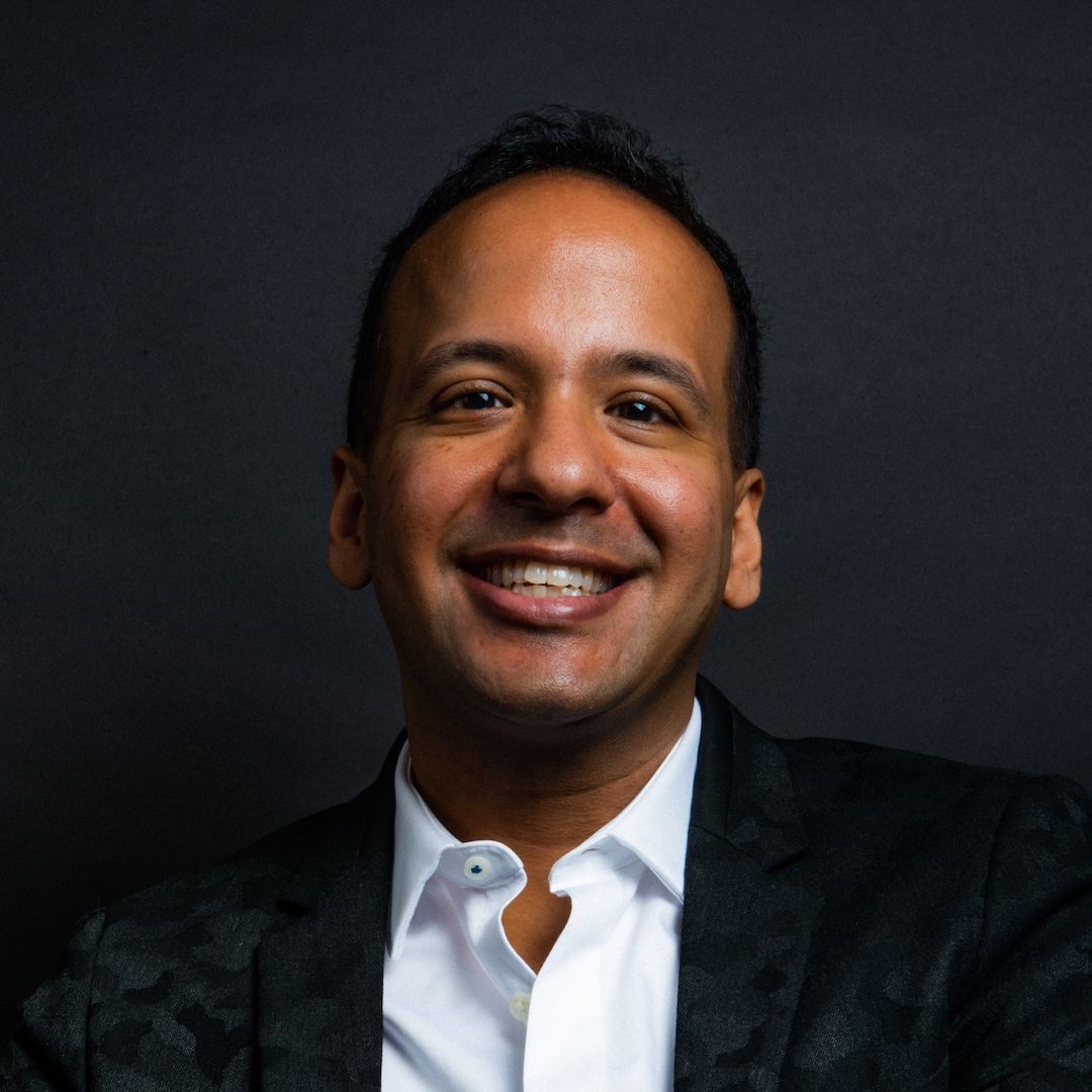 Headshot of keynote speaker, Prasanna Ranganathan, smiling in a black jacket and white shirt, in front of a dark background.