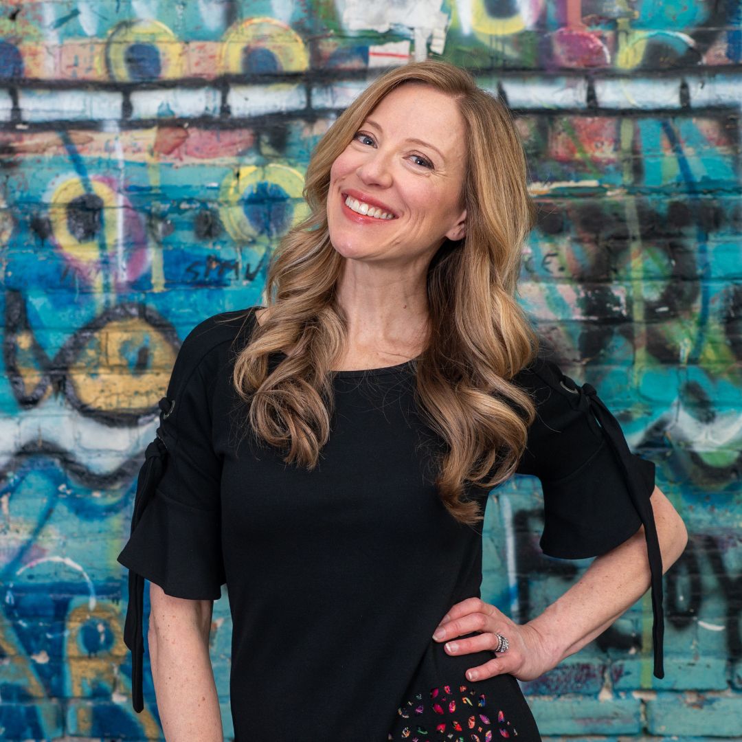 Sarah McVanel. Profile photo of Sarah McVanel, posing with her hand on her hip, smiling, standing in front of a brightly coloured painted brick wall.