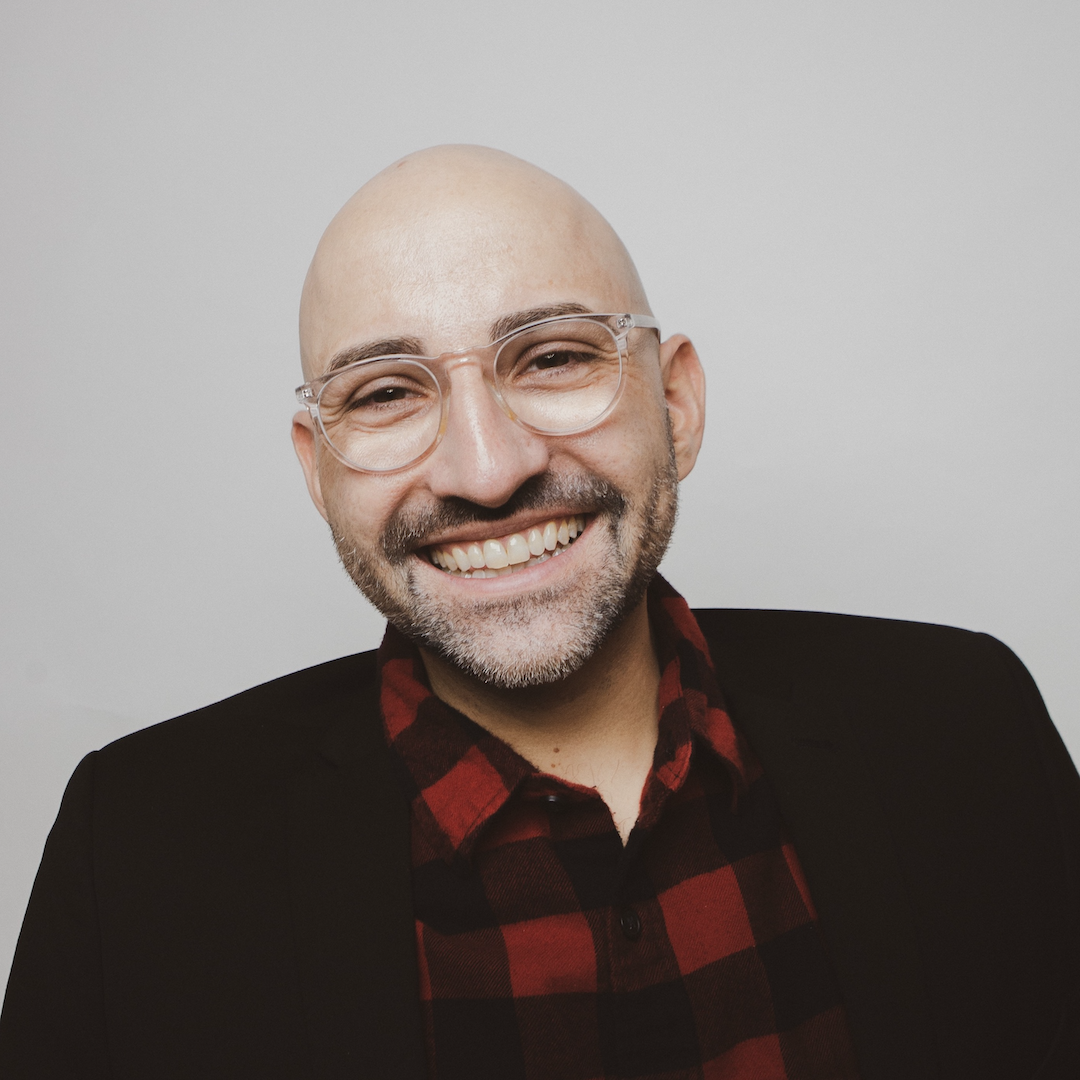 Spencer West. Headshot of Spencer West wearing a black jacket of a red plaid shirt. He is smiling and posing in front of a plain grey background.