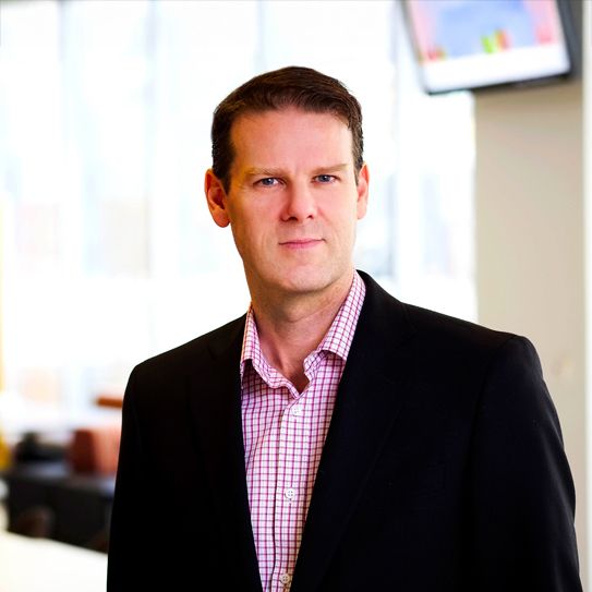 Ted Graham. Headshot of Ted Graham wearing a black jacket over a pink checkered shirt. He is standing in a bright room.