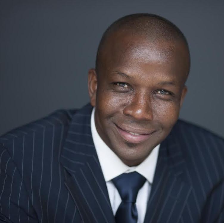 Headshot of Donovan Bailey, black male smiling at the camera, wearing a black striped suit and tie in front of a dark grey backdrop