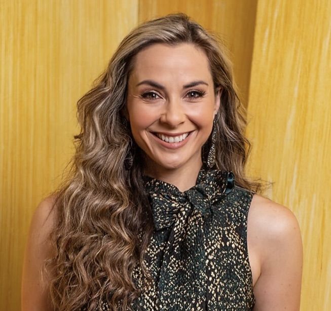 Andi Petrillo. Headshot of Andi Petrillo wearing a black and gold sleeveless top. She is smiling and posing in front of a yellow wall.