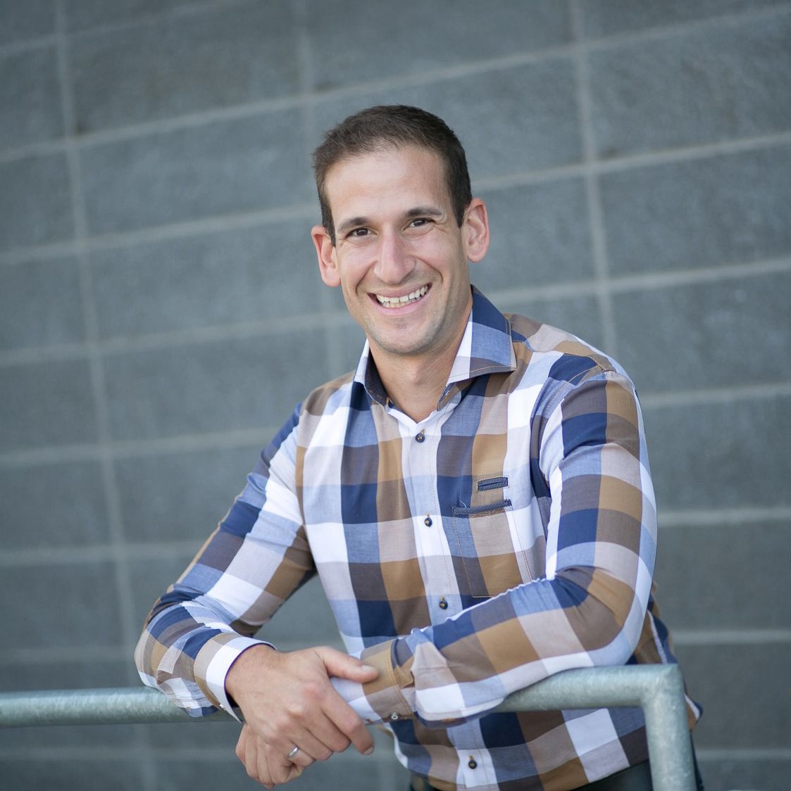 Ramy Nassar. Headshot of Ramy Nassar wearing a blue, brown and white checkered shirt. He is leaning on a metal railing and standing in front of a grey brick wall.