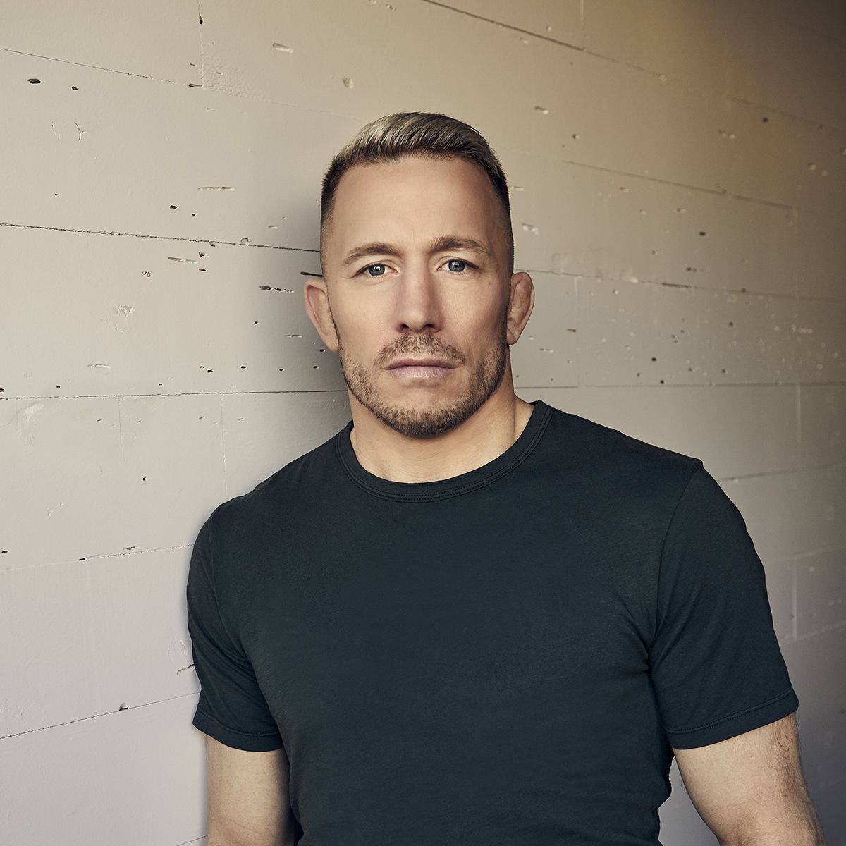 Georges St Pierre. Headshot of Geroges St Pierre wearing a black T-shirt, standing in front of a white brick wall.