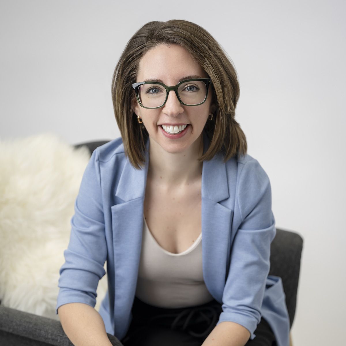 Ashley Margeson. Headshot of Ashley Margeson, wearing a light blue jacket over a white top, sitting sideways on a grey armchair. She is wearing glasses and smiling.