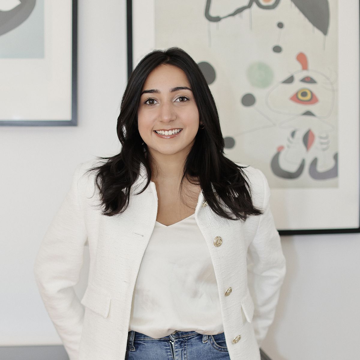 Inana Lalani. headshot of Inana Lalani wearing a white jacket over a white top. She is standing in front of a white wall with a framed piece of abstract art. She is smiling and has her hands in her pockets.