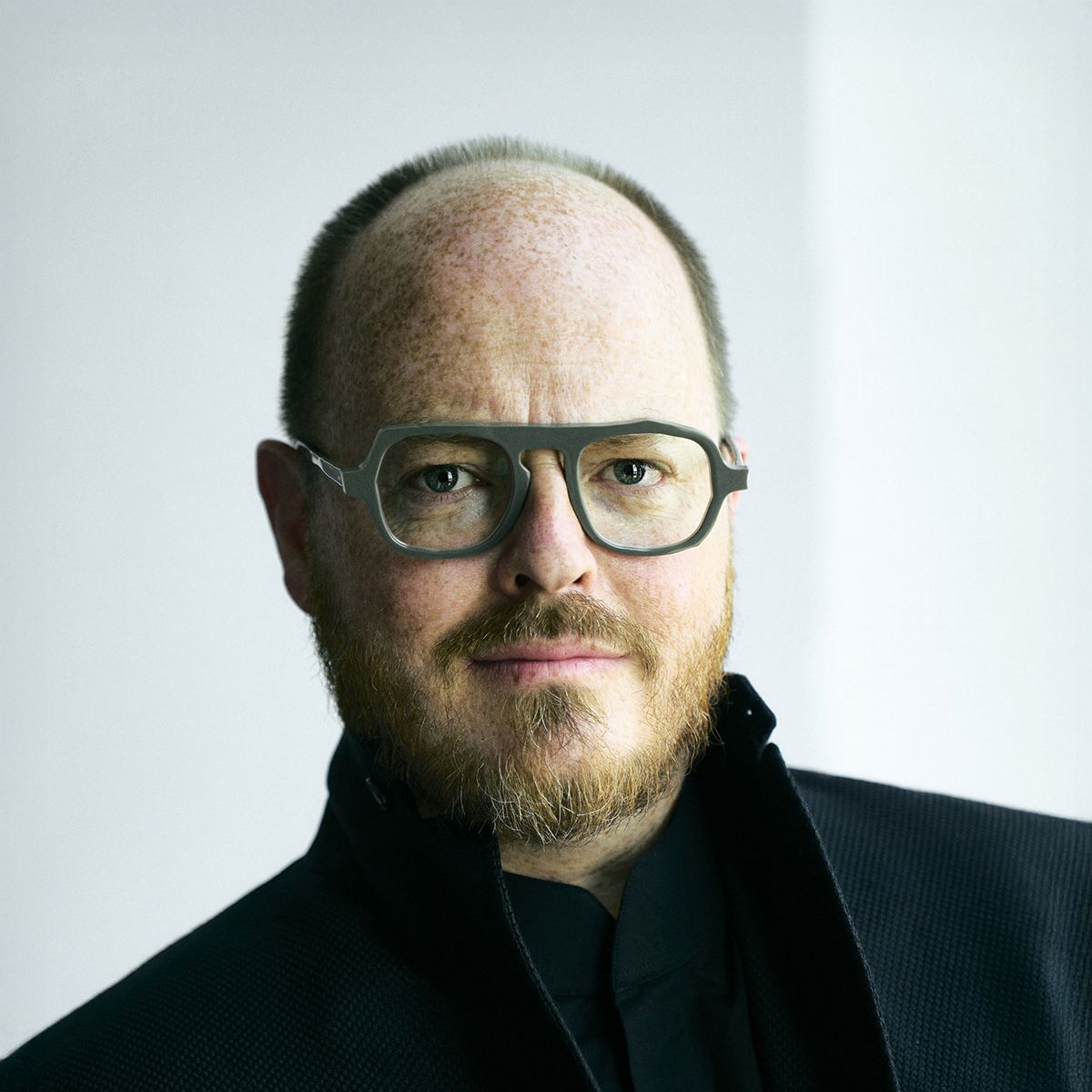 Jonathan Brill. Headshot of Jonathan Brill wearing a black jacket over a black shirt. He is wearing glasses and standing in front of a light grey background.