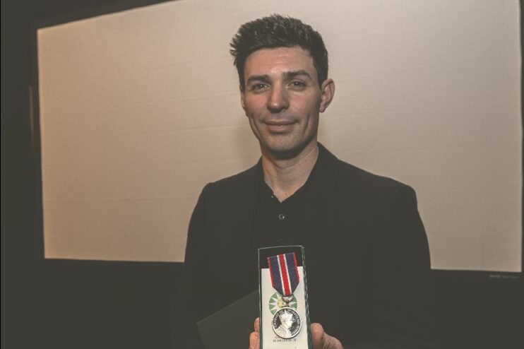 Hockey legend, Carey Price, wearing a black blazer and smiling as he holds up the King Charles III Coronation medal towards the camera.