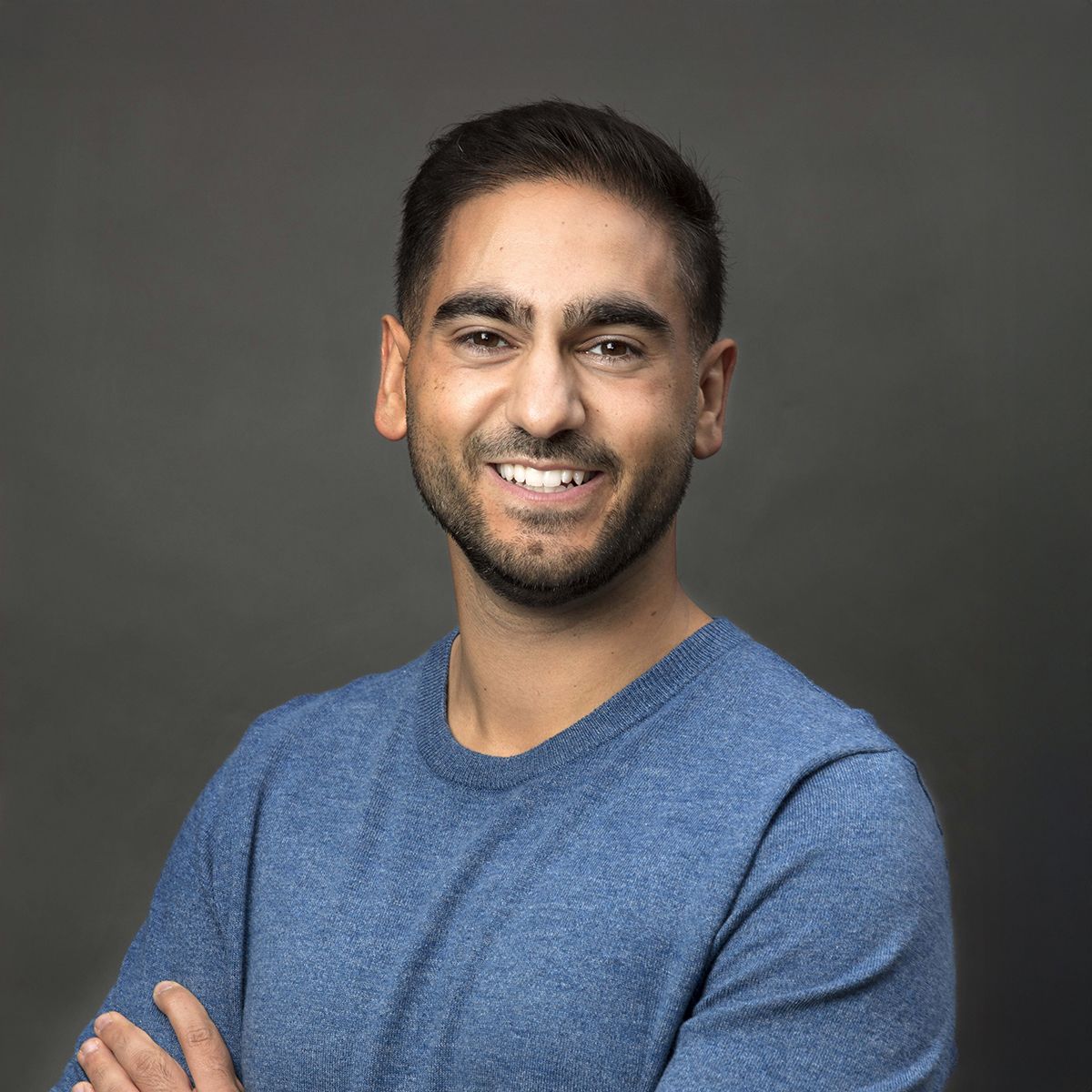 Alex Banayan. Headshot of Alex Banayan wearing a blue top and standing in front of a dark grey background. He is smiling and is crossing his arms.
