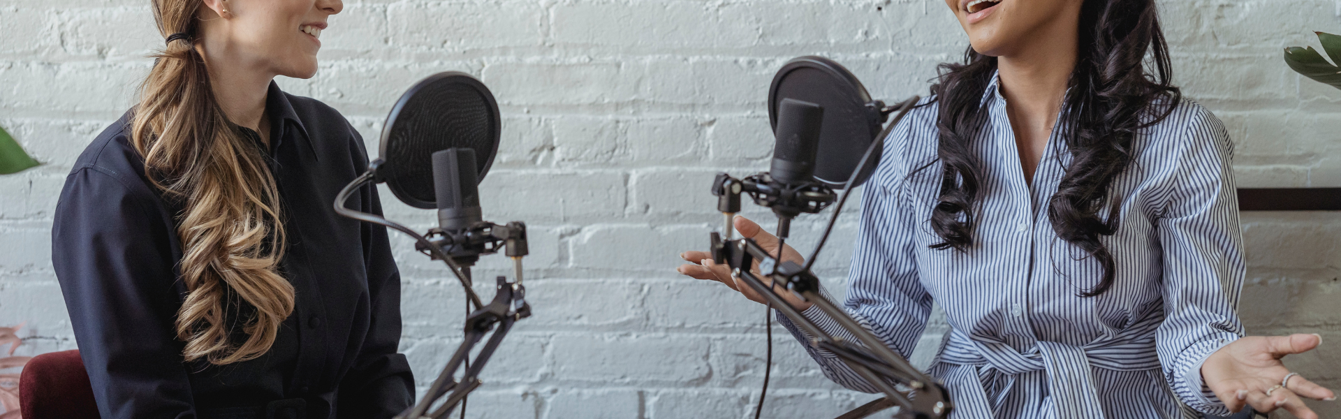 Banner image of two women sitting down at a desk in front of two microphones having a conversation while filming a video or audio podcast.