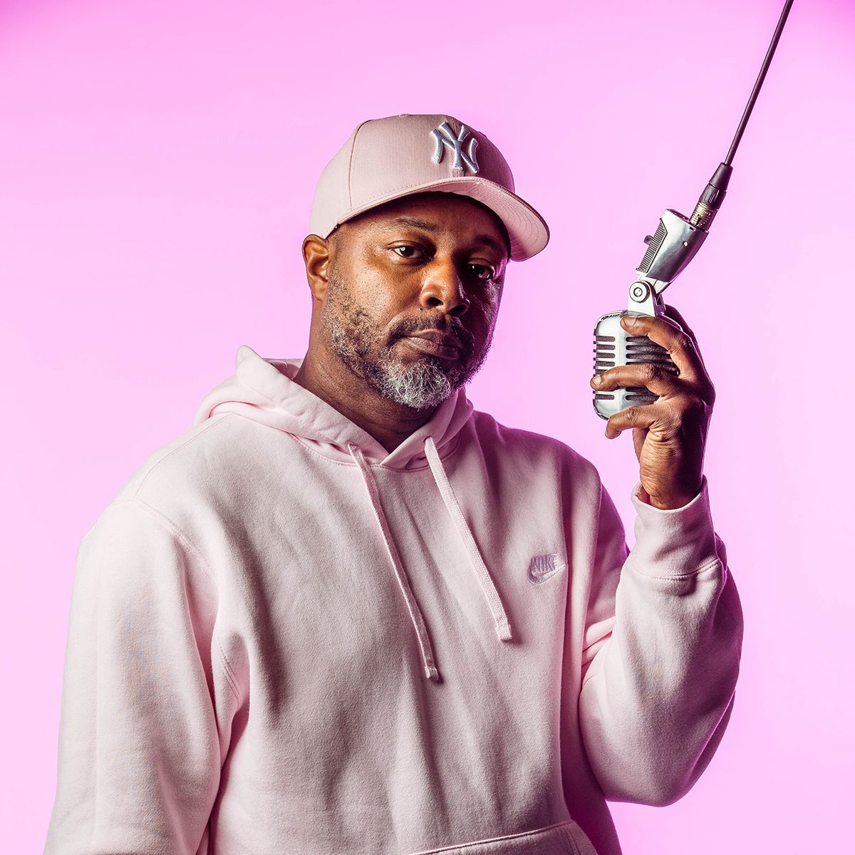 Dwayne Morgan. Headshot of Dwayne Morgan wearing a light pink sweatshirt and cap. He is posing with a microphone and standing in front of a bright pink background.