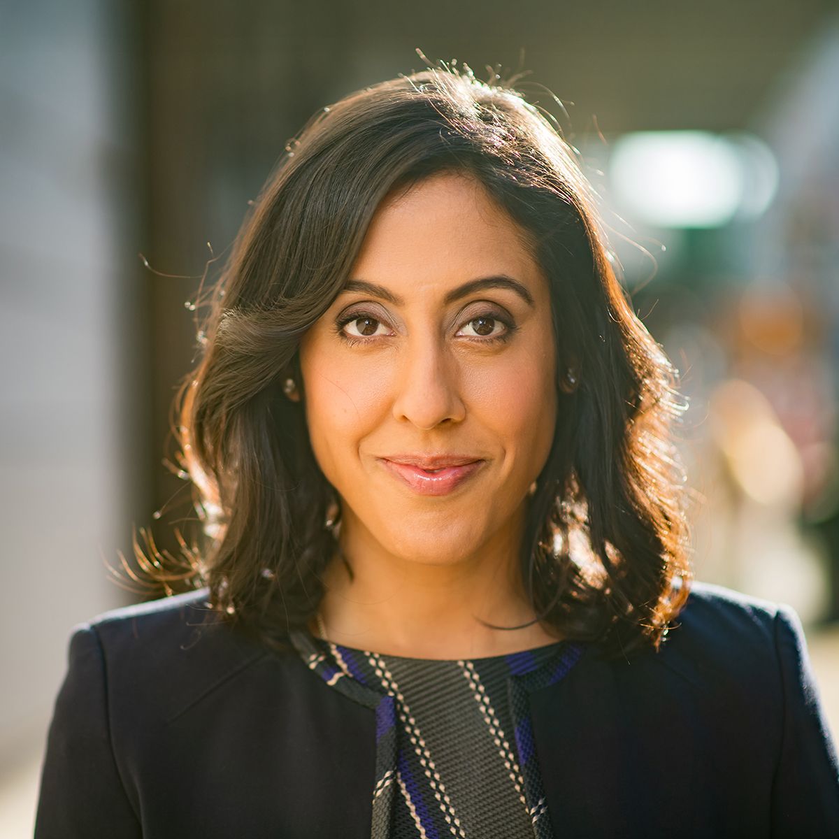 Erica Dhawan. Headshot of Erica Dhawan wearing a black top. She is standing outside and is backlit with a blurred background.