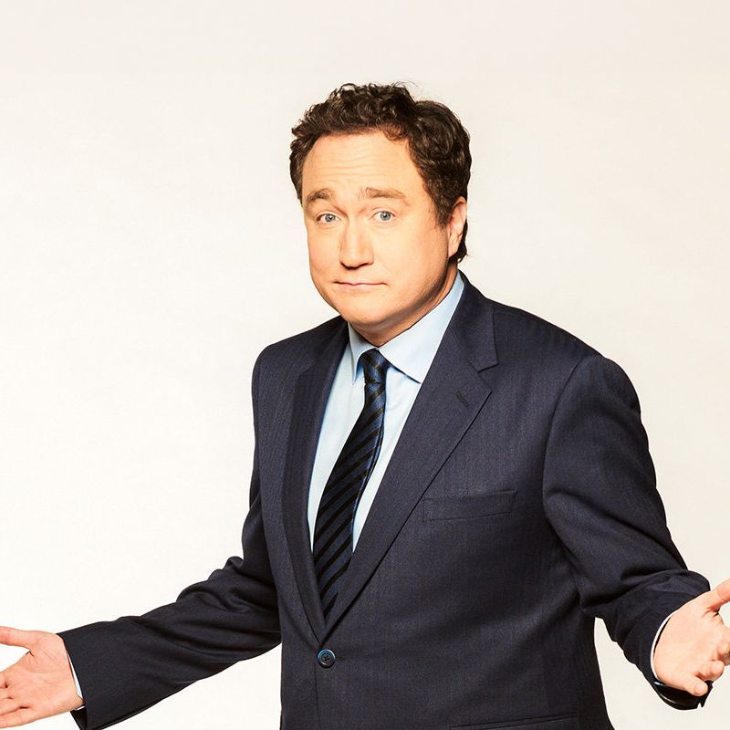 Mark Critch. Headshot of Mark Critch wearing a black suit and tie. He is standing with his arms apart in front of a white background.