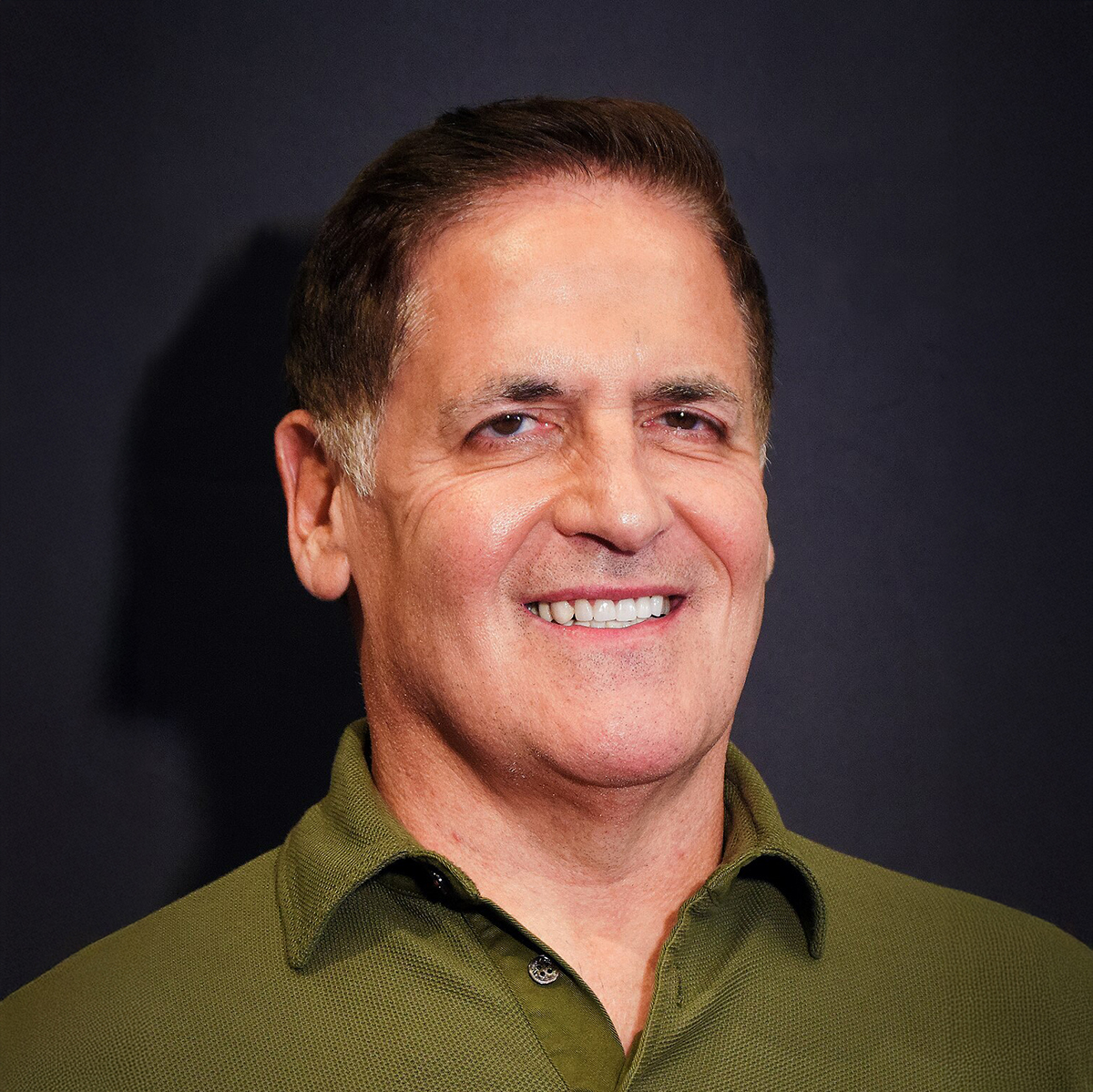 Mark Cuban. Headshot of Mark Cuban wearing a dark green collared top and posing in front of a dark grey background.