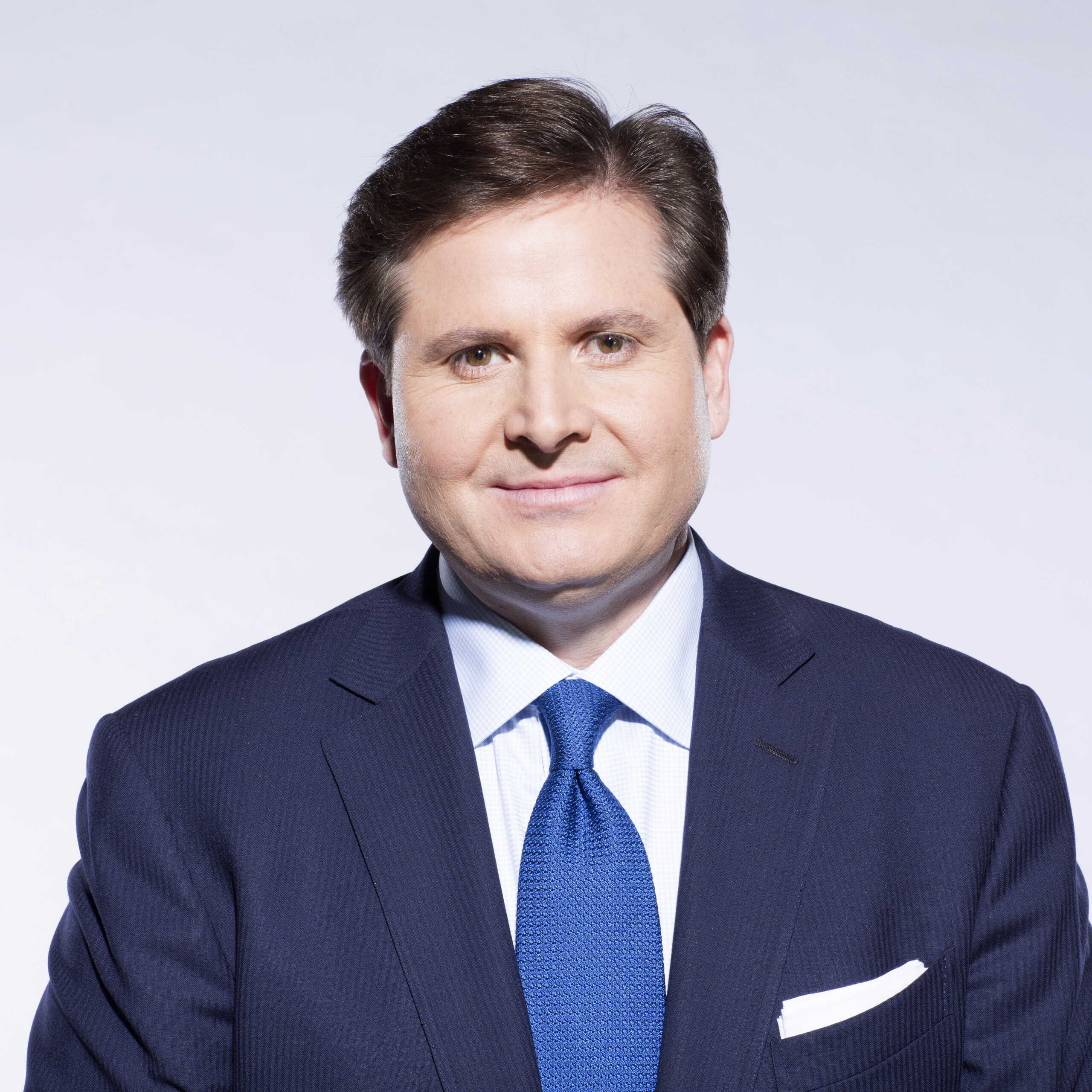 Matt Devlin. Headshot of Matt Devlin wearing a dark navy suit over a white shirt with a blue tie. He is posing in front of a grey gradient background.