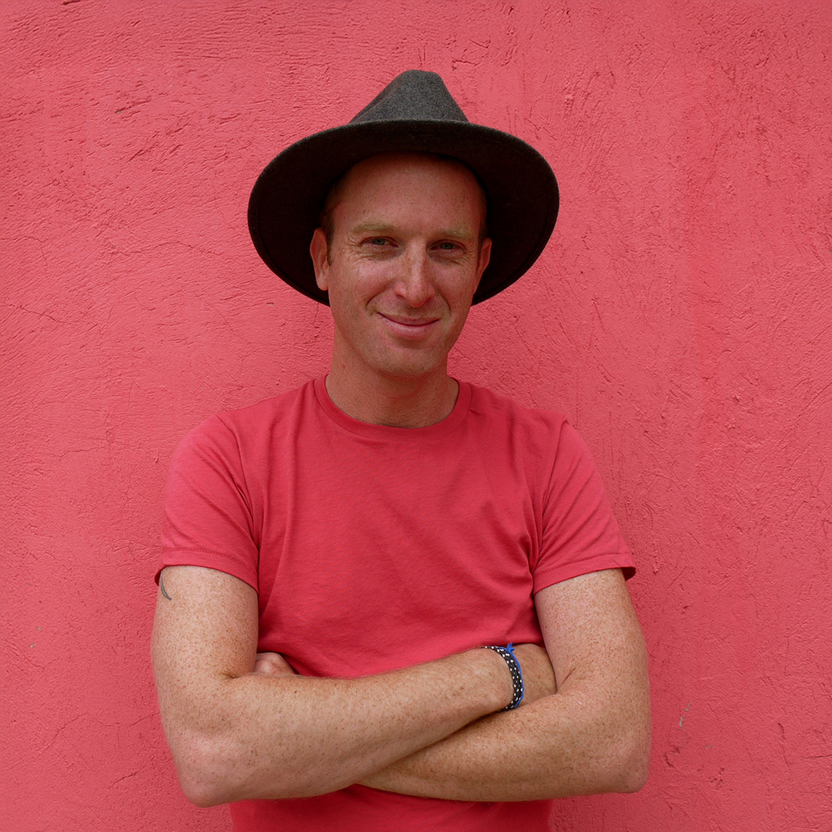 Robin Esrock. Headshot of Robin Esrock. He is wearing a red T-shirt and a black wide-rimmed had. He is crossing his arms and standing in front of a red wall.