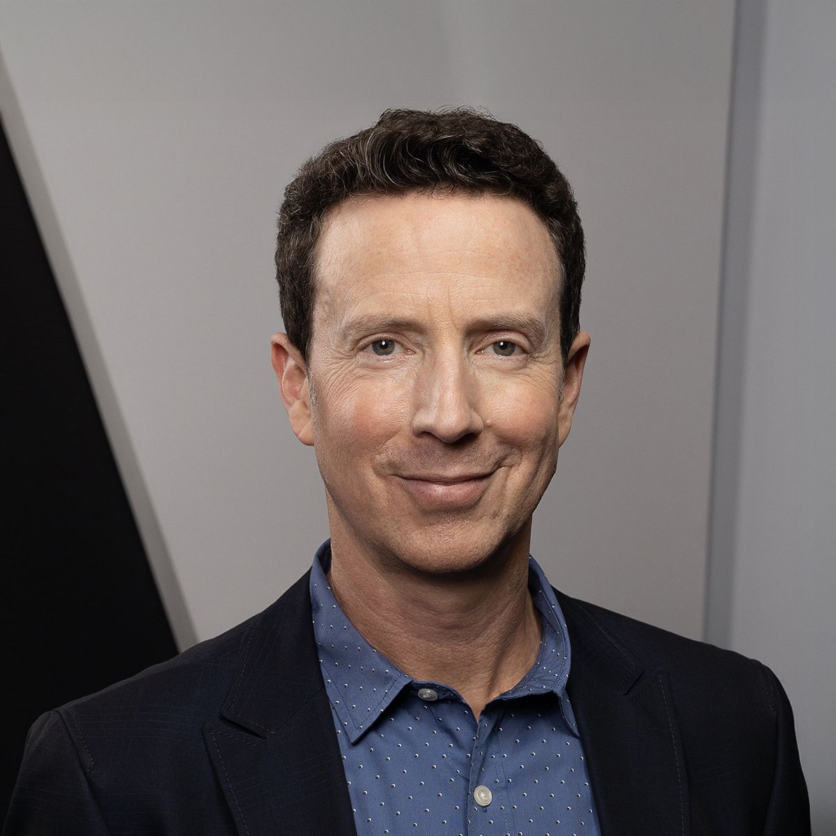 Adam Growe. Headshot of Adam Growe hearing a black suit jacket over a blue collared shirt. He is standing in front of a grey wall.