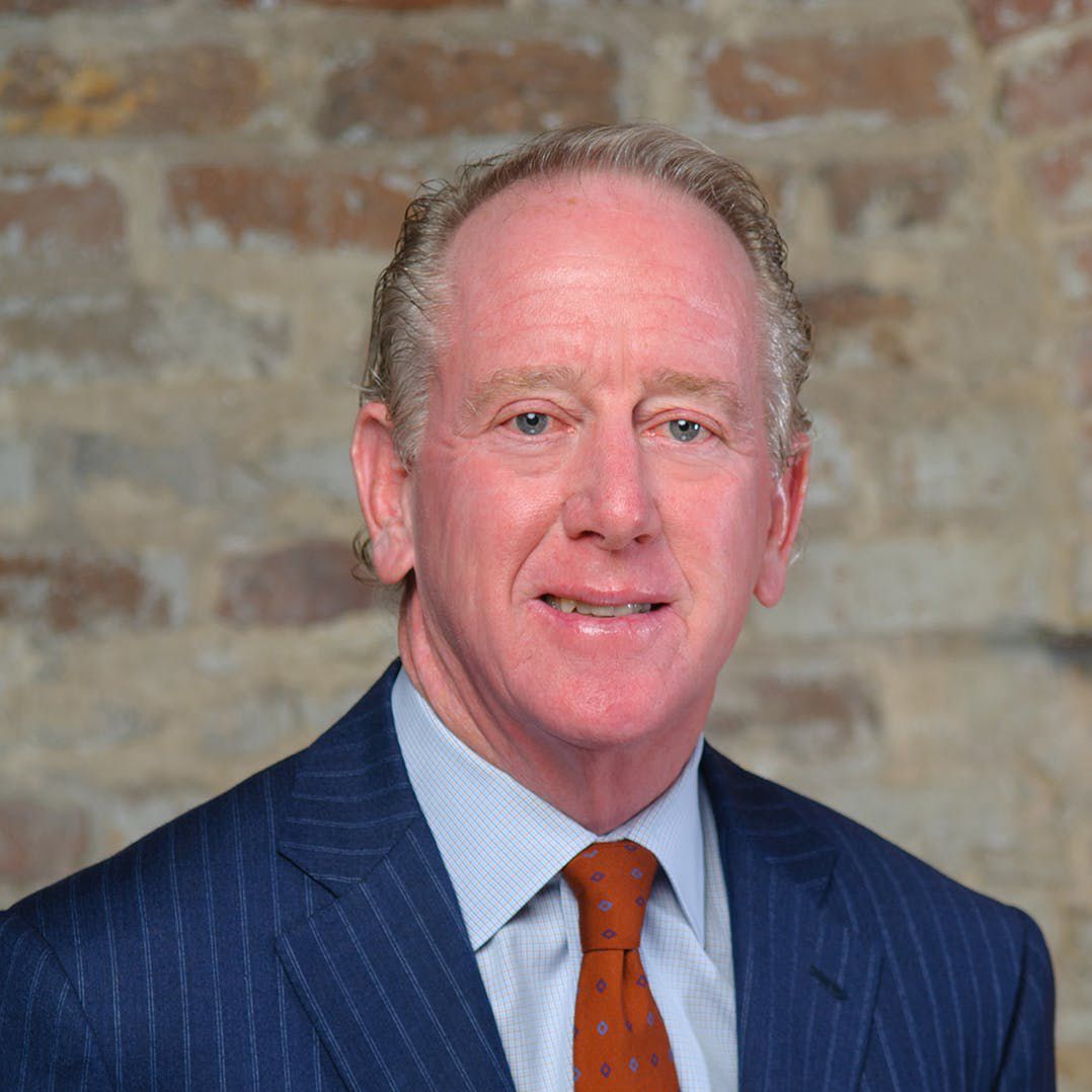Archie Manning. Headshot of Archie Manning wearing a dark blue suit, light blue shirt and a red tie. He is posing in front of a brick textured background.