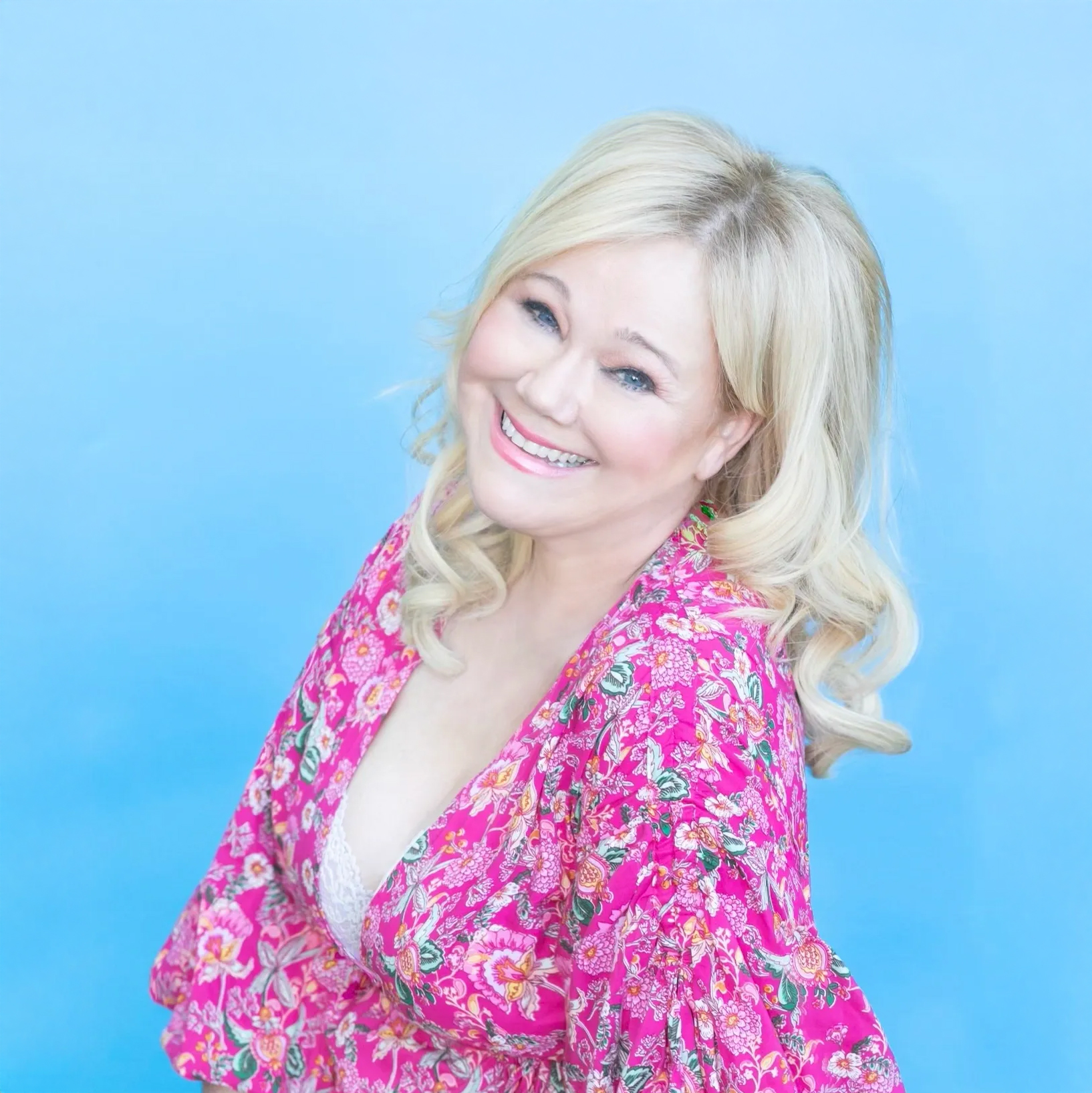 Caroline Rhea. Headshot of Caroline Rhea wearing a pink flowered top. She is posing in front of a bright blue backdrop.