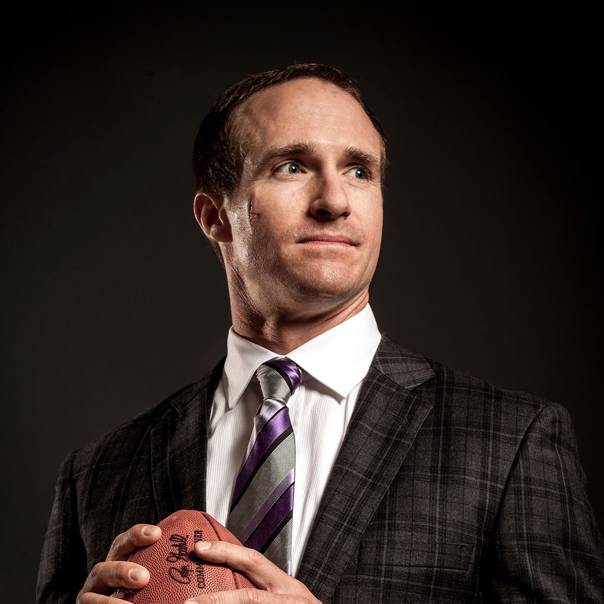 Drew Brees. Headshot of Drew Brees wearing a dark plaid suit, white shirt and tie. He is holding a football and looking off to the side. He's posing in front of a dark backgroud.