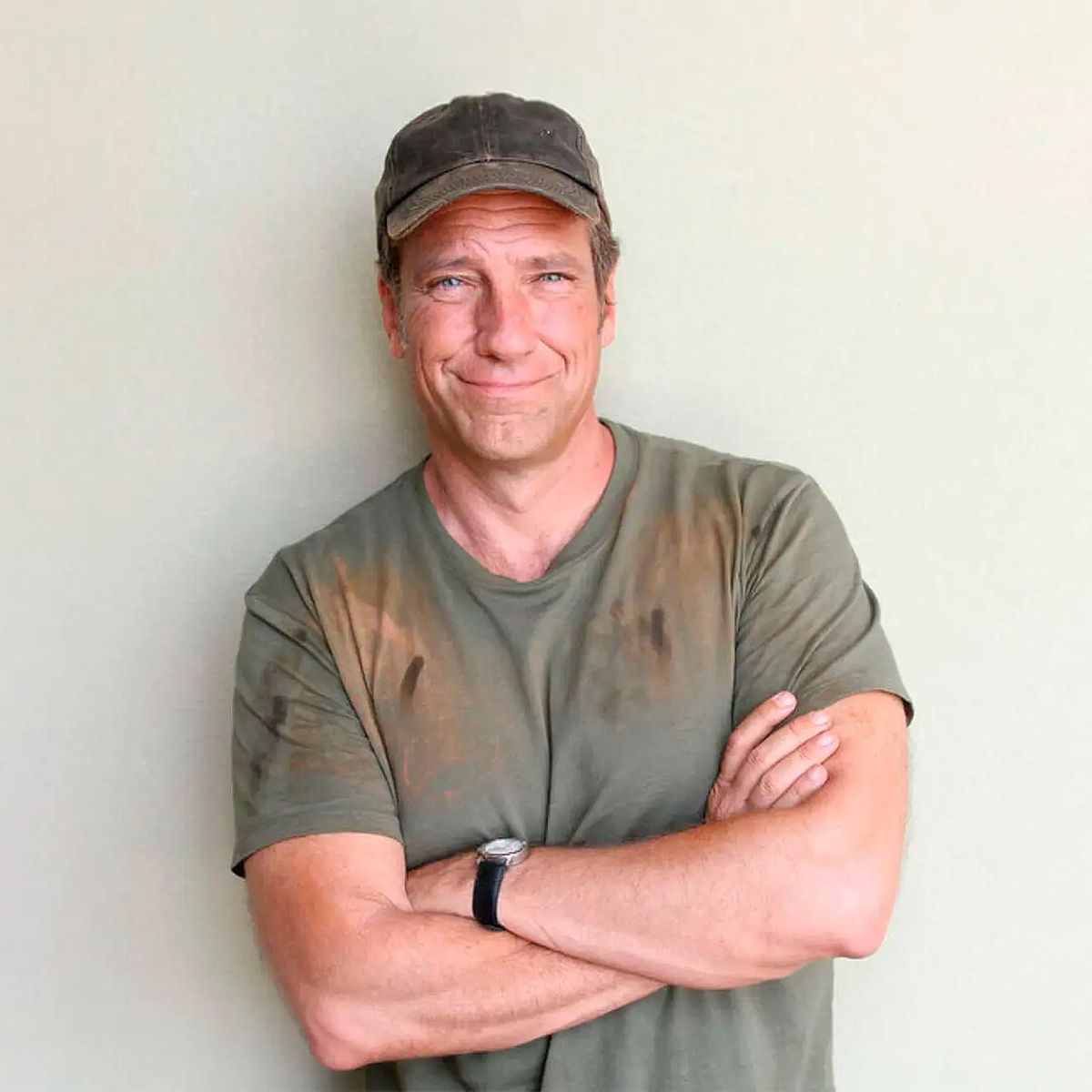 Mike Rowe. Headshot of Mike Rowe wearing a roughed up t-shirt and cap. He's standing with his arms crossed and leaning back on a white wall.