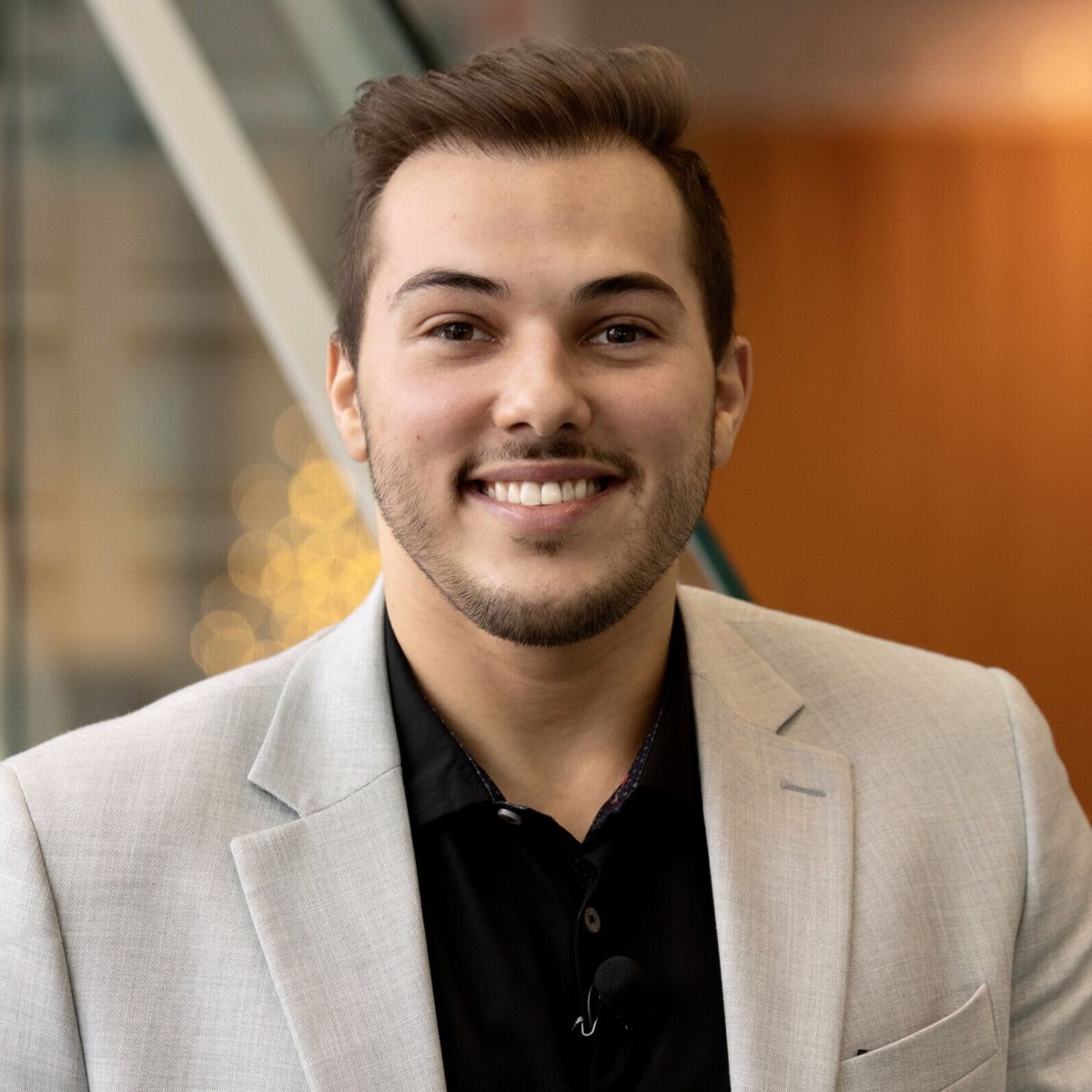 Headshot of Dr. Kaleb Dahlgren, facing the camera and smiling. He is wearing a black shirt and light grey blazer. The backdrop the blurred interior of a building including an orange-coloured wall and a staircase to the left of kaleb.