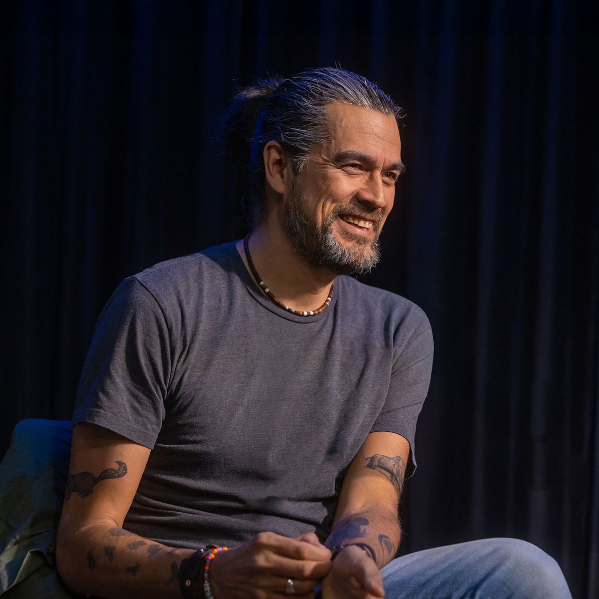 David A Robertson. Headshot of David Robertson wearing a dark grey t-shirt, sitting on stage in front of a dark background.
