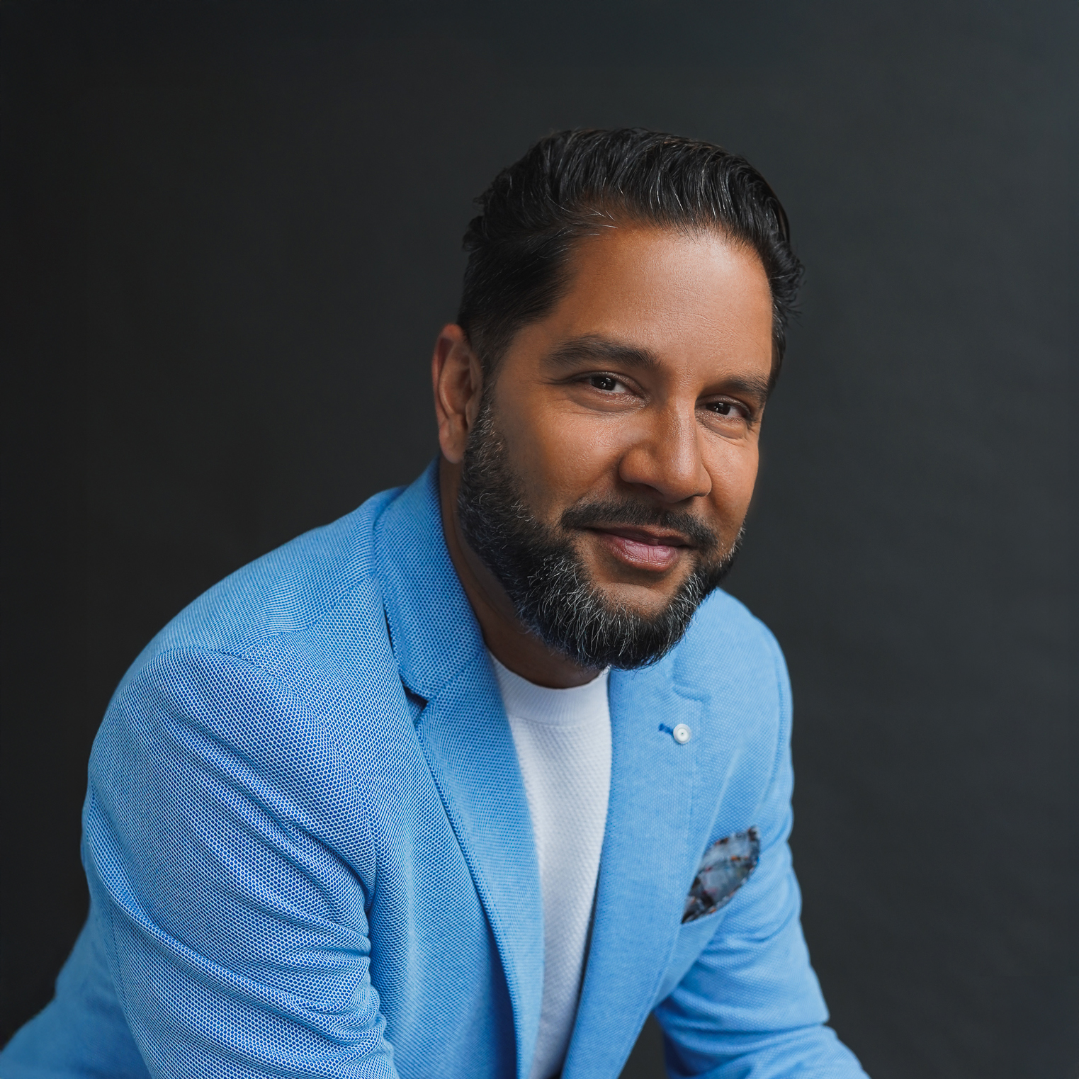 Devo Brown. Headshot of Devo Brown wearing a bright blue suit jacket over a white shirt. He is leaning forward and posing in front of a dark background.