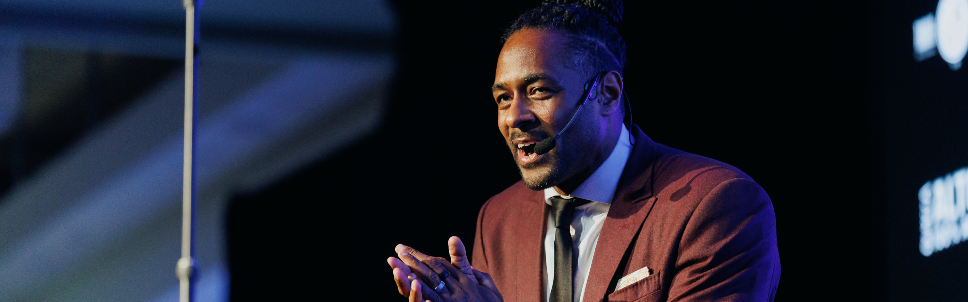Jesse Lipscombe onstage hosting an event. He is wearing a maroon suit.