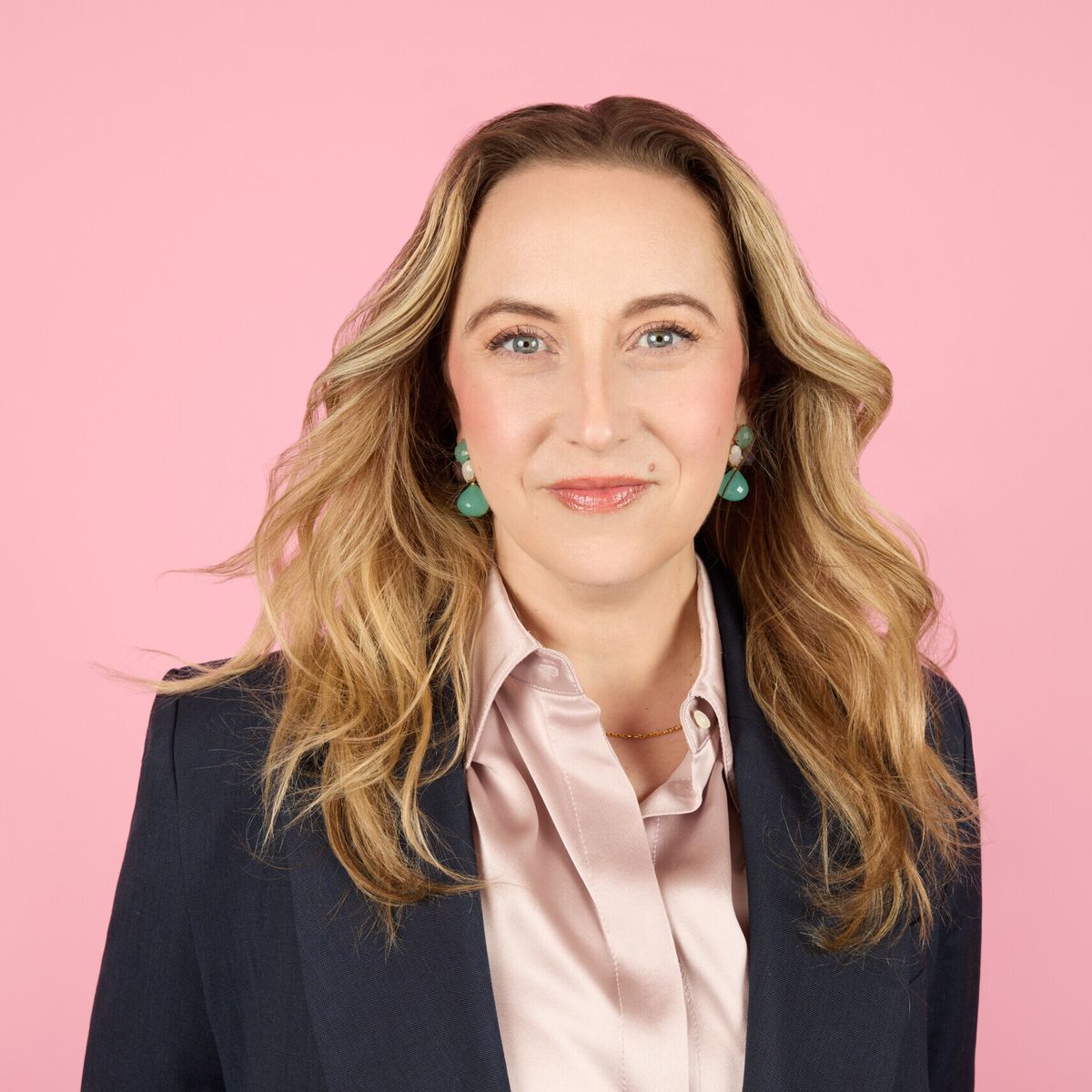 Headshot of Jodi Kovitz in front of a solid pink backdrop. She is smiling and wearing a black blazer jacket over a satin blush-coloured button-up shirt.