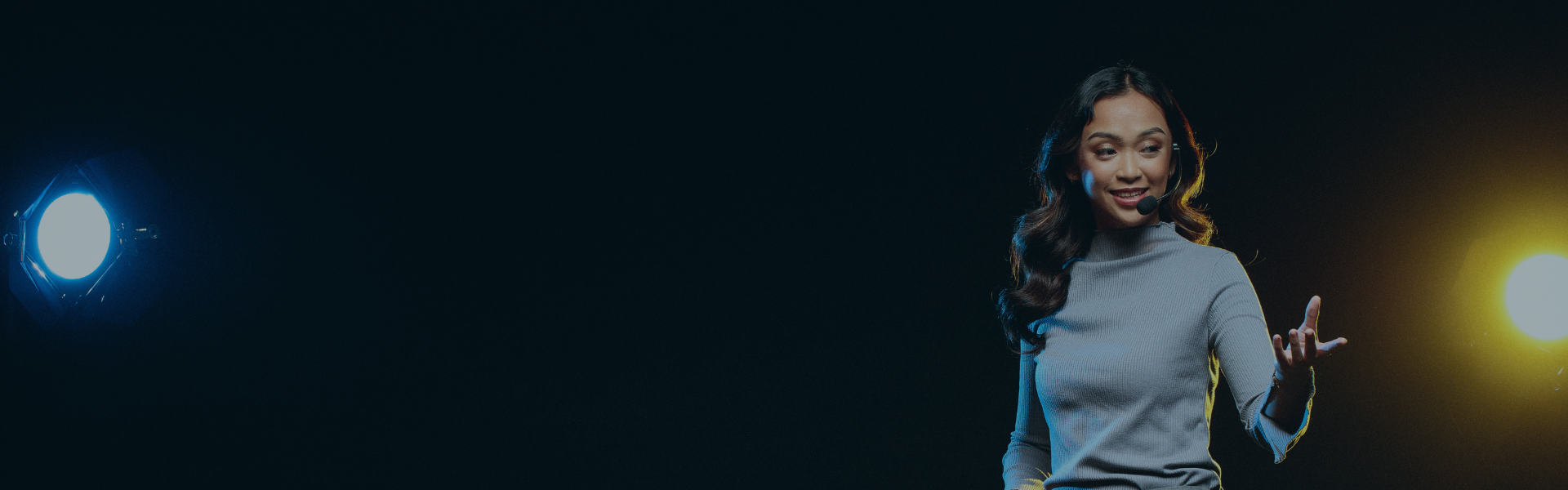 A female keynote speaker on stage delivering a speech. The background is solid black with 2 stage lights shining on either side of the subject.
