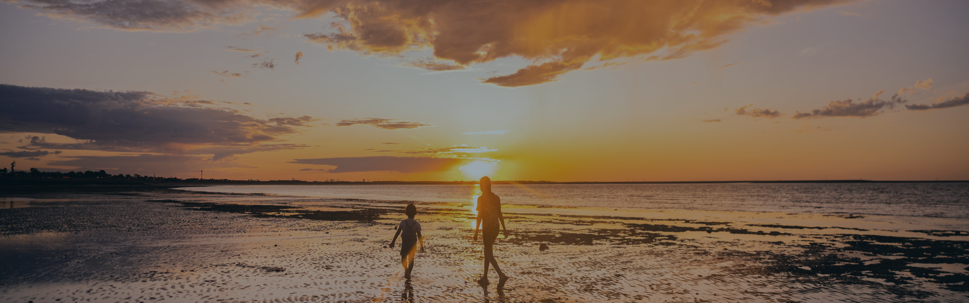 Two people walking on a beach next to the shoreline and as the sun sets in the background creating an orange-coloured sky.