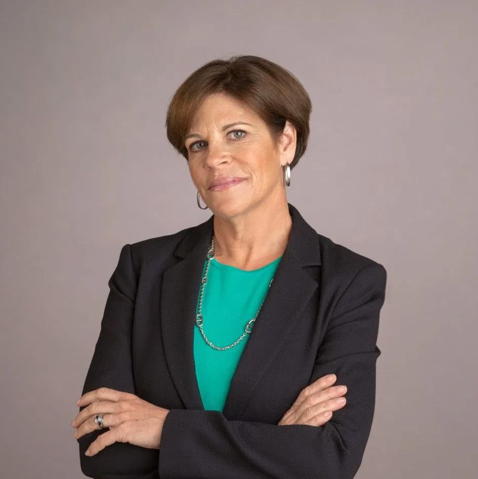 Michelle Stacey. Headshot of Michelle Stacey wearing a bloack blazer over a green top. She's posing with her arms crossed in front of a grey background.