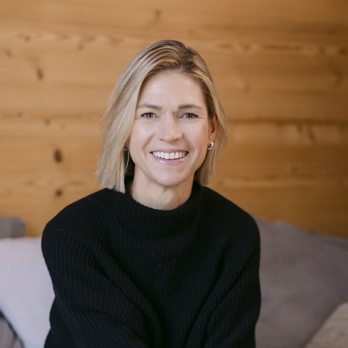 Headshot of speaker, Stephanie Case, wearing a black long-sleeve shirt and white pants. She is sitting on a grey couch, and smiling at the camera.
