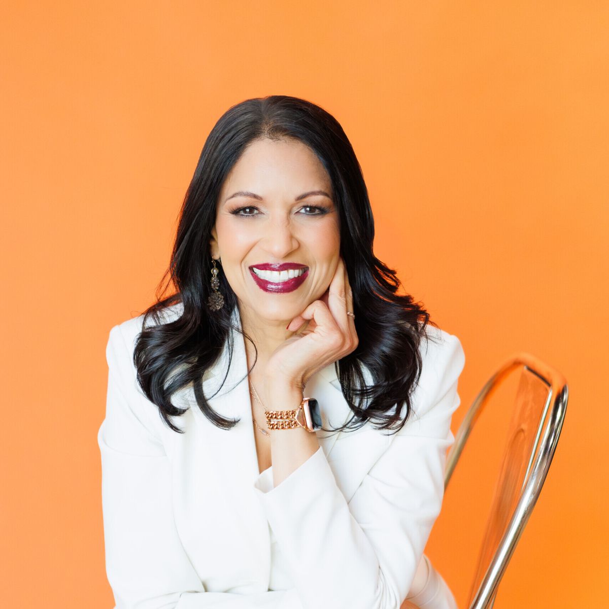 Rashmi Airan, keynote speaker, professional headshot. She is wearing an all white pant suit and is sitting on a chair in front of a solid orange backdrop.