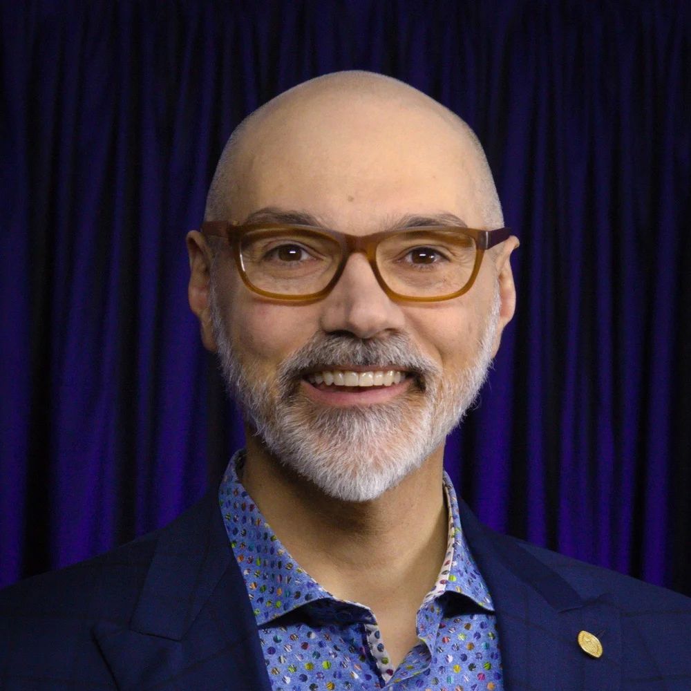 Dan Trommater. Headshot of Dan Trommater wearing a blue jacket over a blue collared shirt. He is posing in front of a dark blue curtain.