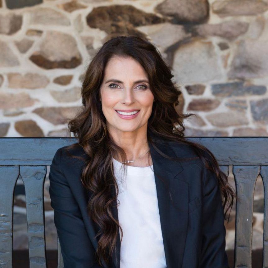 Ryan Haddon. Headshot of Ryan Haddon wearing a navy jacket over a white top. She is sitting outside on a bench in front of a stone wall.