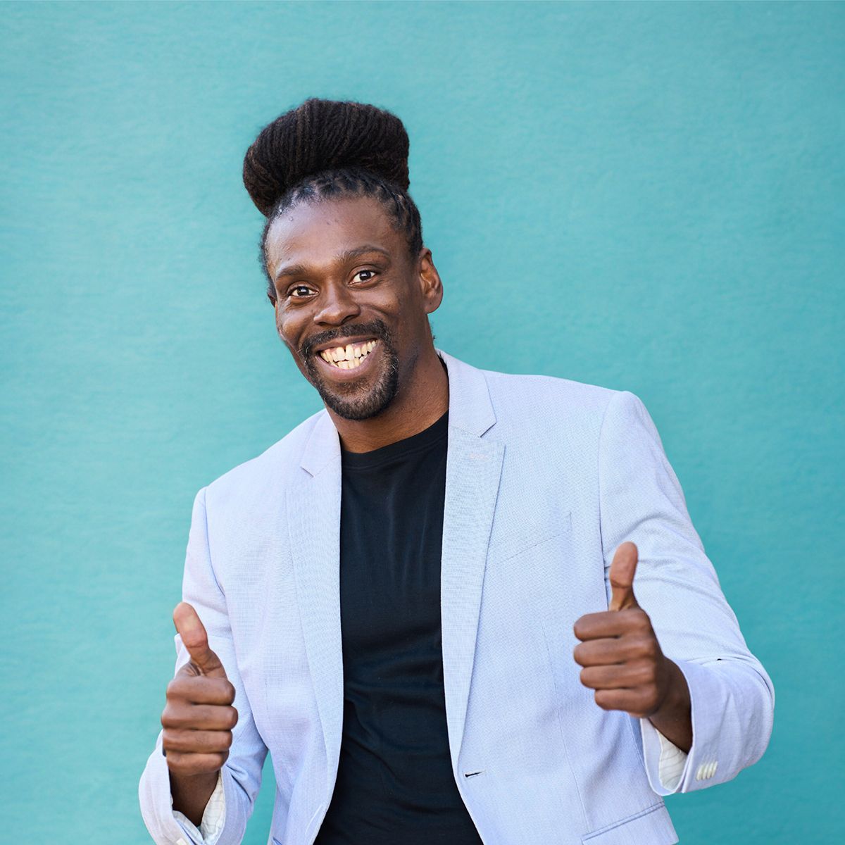Cam F. Awesome. Headshot of Cam F. Awesome wearing a white jacket over a black top. He's posing with his thumbs up in front of a teal wall.