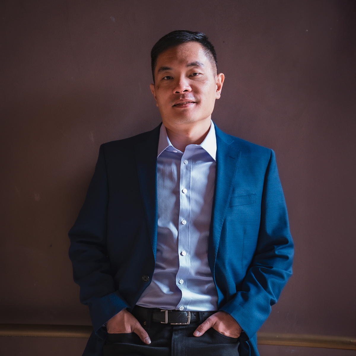 Jia Jiang. Headshot of Jia Jiang wearing a dark blue suit over a light blue shirt. He's posing with his hands in his pockets in front of a dark brown background.