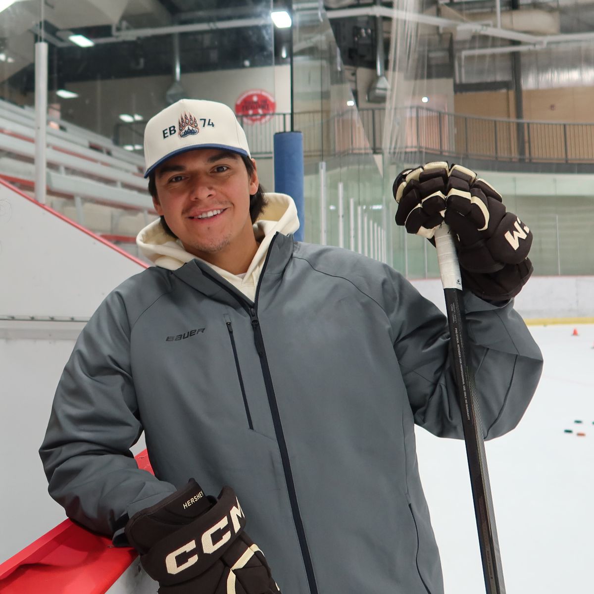 Ethan Bear. Headshot of Ethan bear posing in a hockey arena wearing a grey jacket, ball cap and hockey gloves.
