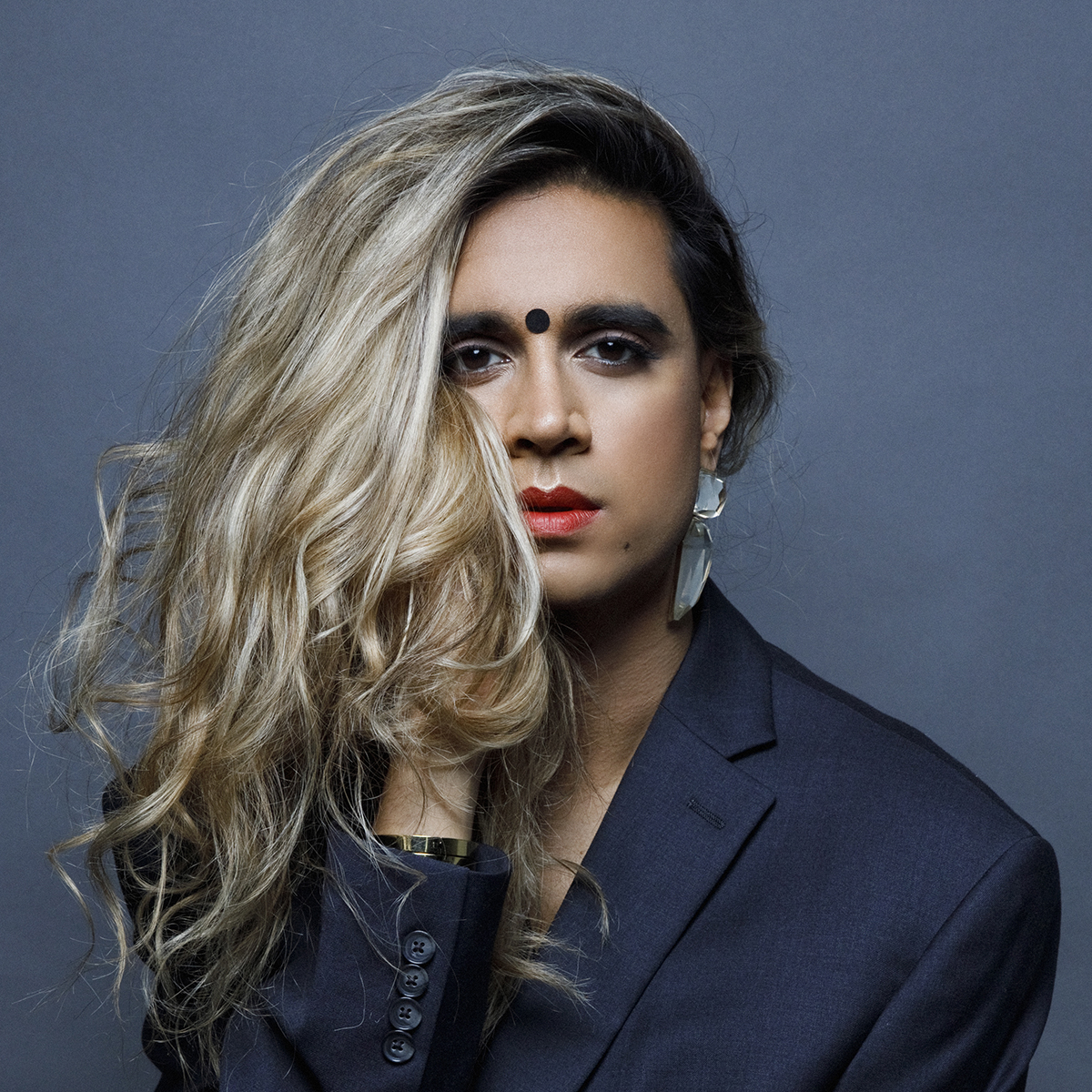 Vivek Shraya. Headshot of Vivek Shraya wearing a dark navy jacket, posing with her hand under her chin, in front of a dark grey background.