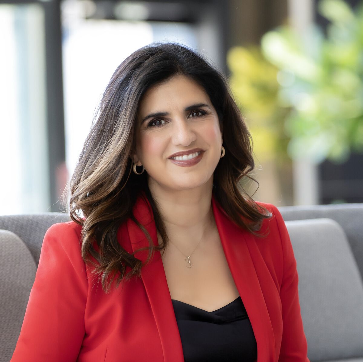Headshot of Fotini Iconomopoulos smiling and sitting on a grey couch. She is wearing a black top underneath a red blazer.