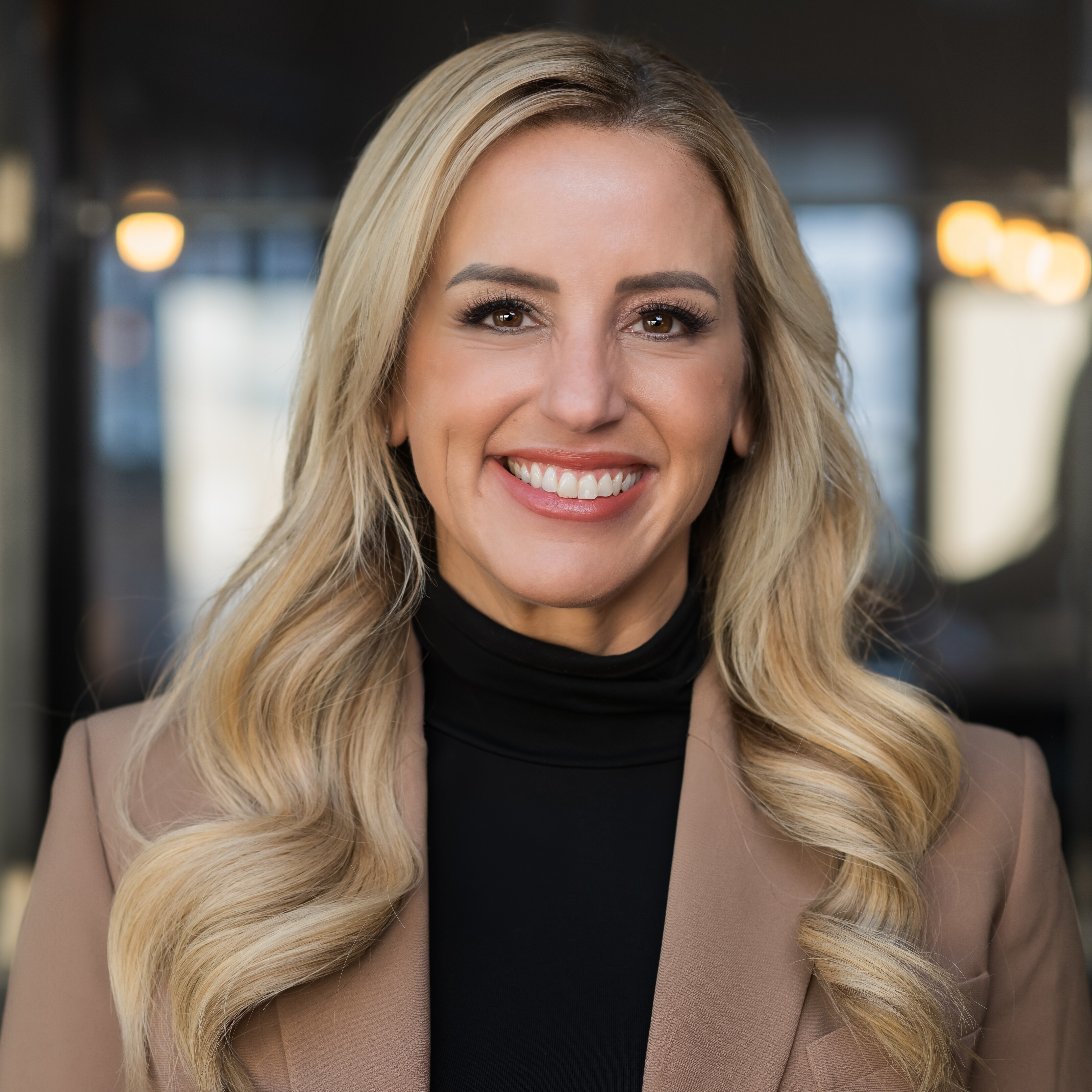 Headshot of keynote speaker, Rachel DeAlto, in front of a blurred background. She is smiling, wearing a black turtle neck shirt underneath a tan blazer jacket.